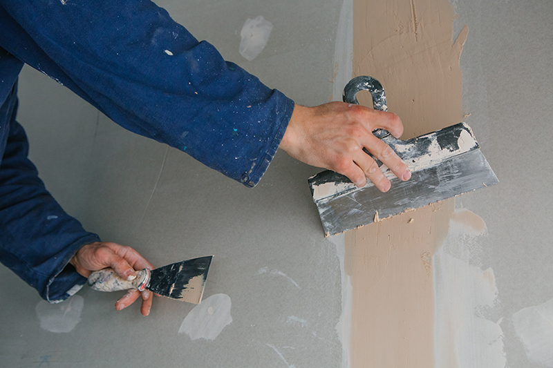 Foto das mãos do profissional gesseiro passando a espátula para gesso na parede